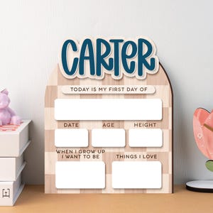 Op de afbeelding: Houten mijlpaalbord met de naam "CARTER" in blauw schrift. Het bord heeft vakken voor "DATUM", "LEEFTIJD" en "LENGTE", samen met vragen voor "ALS IK GROOT BEN WIL IK WORDEN" en "Dingen die ik leuk vind". De achtergrond heeft een lichtbruin gestreept patroon.
