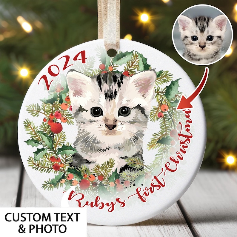 May include: A round Christmas ornament featuring a kitten's portrait, surrounded by a wreath of greenery and red berries. The ornament includes the text "2024" and "Ruby's first Christmas". A small photo of the kitten is in the upper right corner.