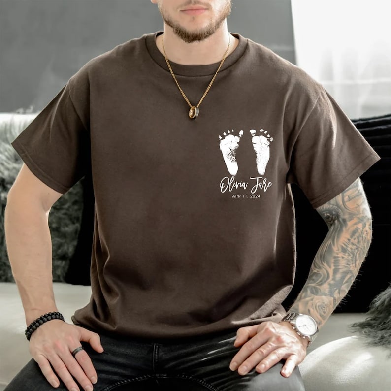 May include: A brown t-shirt with a white imprint of two baby footprints and the text "Olivia Jane APR 11, 2024".