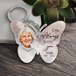 May include: A silver butterfly-shaped keychain with a watercolor background. The keychain features a photo of a woman and the text "I AM Always WITH you MOM 1947-2025".