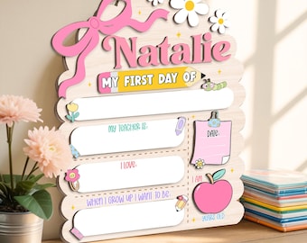 Puede incluir: Letrero de madera con el nombre "Natalie" en rosa, con un lazo rosa, margaritas y el texto "MY FIRST DAY OF". Incluye secciones para rellenar para el profesor, los amores y las aspiraciones futuras, además de un gráfico de manzana y "YEARS OLD."