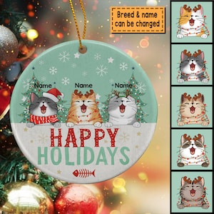 May include: A round ceramic ornament with a green background and a white border. It features three cats wearing Santa hats and reindeer antlers, with the text "Happy Holidays" in red and white letters. The ornament is decorated with snowflakes and a fishbone.