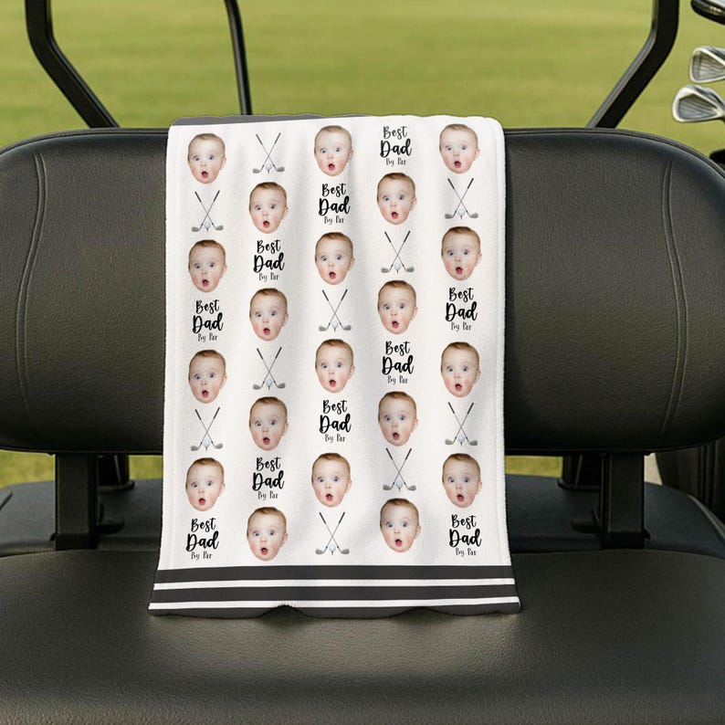 May include: White golf towel featuring a repeating pattern of baby faces, golf clubs, and the text "Best Dad By Far." The towel has a black and white striped border and is on a golf cart.