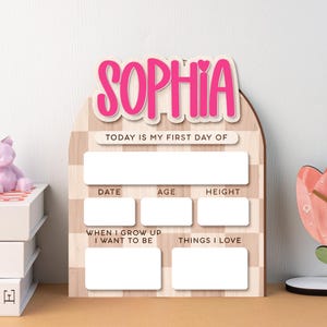 Op de afbeelding: Houten mijlpaalbord met de naam "SOPHIA" in roze letters. Het bord heeft vakken voor "DATE", "AGE", "HEIGHT", "WHEN I GROW UP I WANT TO BE" en "THINGS I LOVE". De tekst "TODAY IS MY FIRST DAY OF" is ook aanwezig.