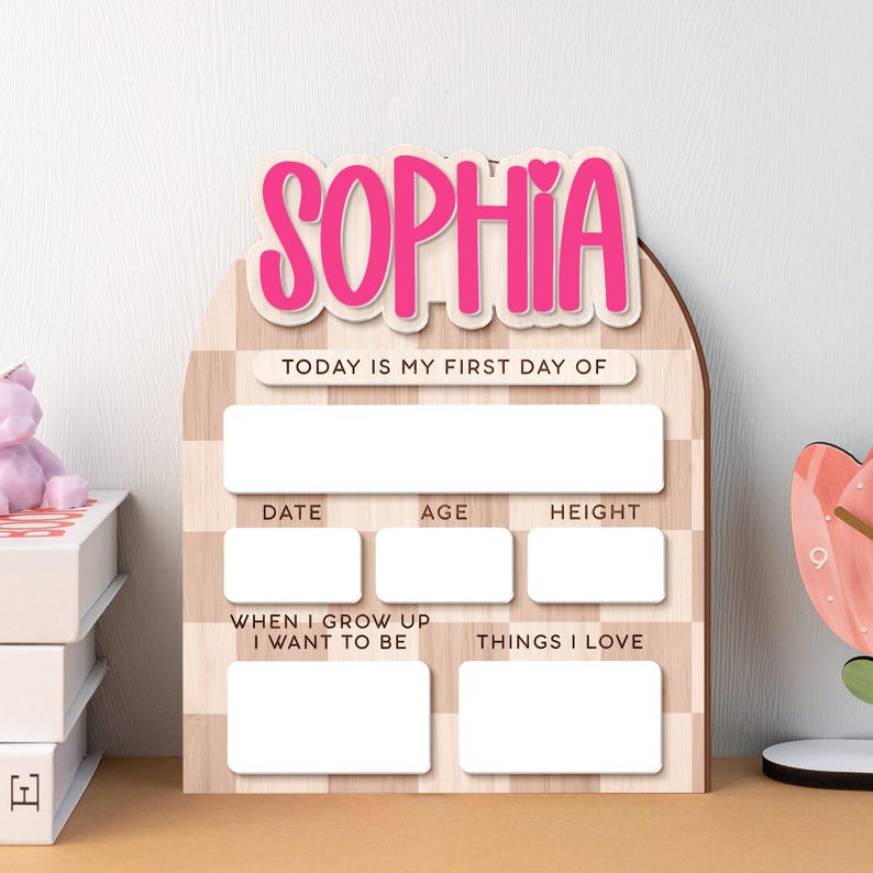 Op de afbeelding: Houten mijlpaalbord met de naam "SOPHIA" in roze letters. Het bord heeft vakken voor "DATE", "AGE", "HEIGHT", "WHEN I GROW UP I WANT TO BE" en "THINGS I LOVE". De tekst "TODAY IS MY FIRST DAY OF" is ook aanwezig.