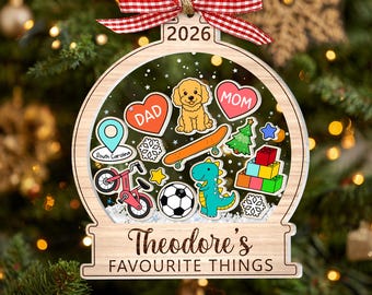May include: A wooden snow globe ornament featuring a red and white gingham bow and the year 2026. The center displays colorful icons including a dog, hearts labeled DAD and MOM, a skateboard, bicycle, soccer ball, dinosaur, and building blocks.