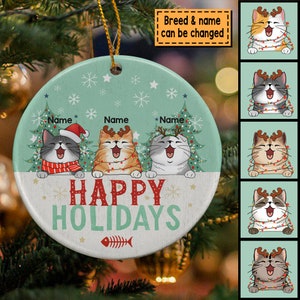 May include: A round, green and white Christmas ornament with three cats wearing Santa hats and reindeer antlers. The cats are sitting in front of a Christmas tree and the ornament says "Happy Holidays".