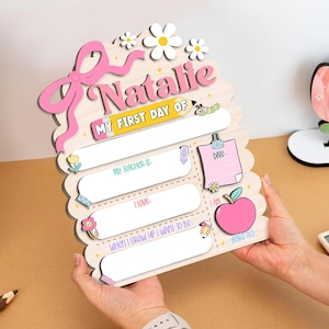 May include: Wooden first day of school sign with the name "Natalie" in pink. Features spaces to write "My Teacher Is", "I Love", and "When I Grow Up I Want To Be". Includes a pink apple, daisies, and the text "My First Day Of".