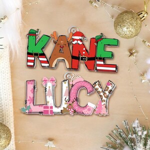 May include: Two Christmas-themed name ornaments. The top ornament spells "KANE" with letters decorated as a Christmas elf, reindeer, and Santa. The bottom ornament spells "LUCY" with pink and patterned letters, and Christmas-themed embellishments.