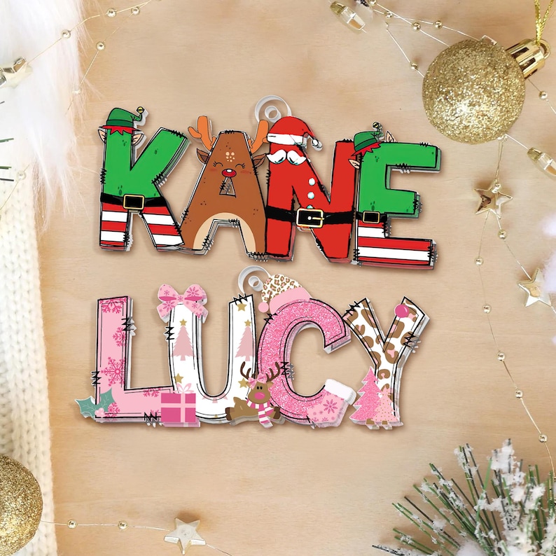 May include: Two Christmas-themed name ornaments. The top ornament spells "KANE" with letters decorated as a Christmas elf, reindeer, and Santa. The bottom ornament spells "LUCY" with pink and patterned letters, and Christmas-themed embellishments.