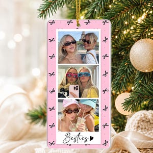 May include: A rectangular pink hanging ornament featuring a repeating black bow pattern and three stacked photographs of two individuals. The bottom of the ornament displays the text Besties in black script with a small black heart icon.