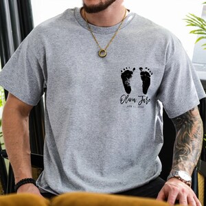 May include: Gray t-shirt with black baby footprints and the text "Olivia Jane APR 11, 2024".