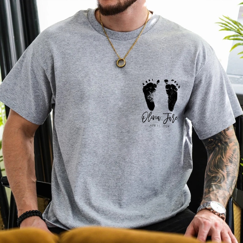 May include: Gray t-shirt with black baby footprints and the text "Olivia Jane APR 11, 2024".