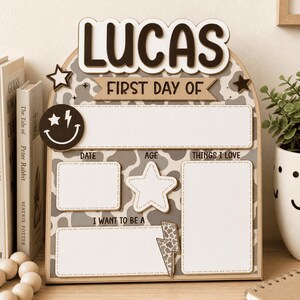 May include: An arched wooden sign with a gray and white cow print background. The text reads LUCAS, FIRST DAY OF, DATE, AGE, THINGS I LOVE, and I WANT TO BE A. It features a smiley face with a lightning bolt eye, two stars, and a lightning bolt.