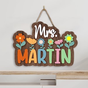May include: A wooden sign with the text "Mrs. Martin" in colorful letters. The sign is decorated with flowers in various colors, including orange, yellow, pink, and blue. The sign is hanging from a rope.