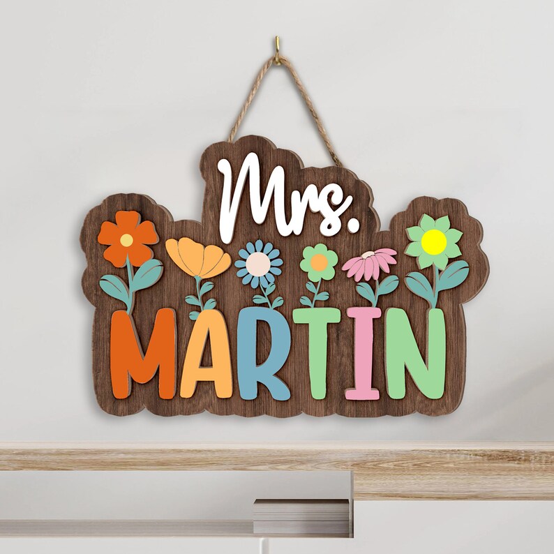 May include: A wooden sign with the text "Mrs. Martin" in colorful letters. The sign is decorated with flowers in various colors, including orange, yellow, pink, and blue. The sign is hanging from a rope.