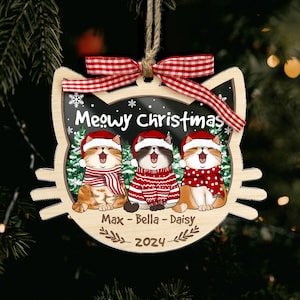 May include: Wooden Christmas ornament shaped like a cat's head, featuring three cartoon cats wearing Santa hats and scarves. The ornament says "Meowy Christmas" and includes the names Max, Bella, and Daisy, with the year 2024.