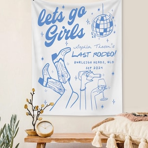 May include: White tapestry with blue text that reads "let's go girls" and "Sophia Thason's Last Rodeo!" with a disco ball graphic and a drawing of two women wearing cowboy boots and holding a bottle of wine and a glass of wine.