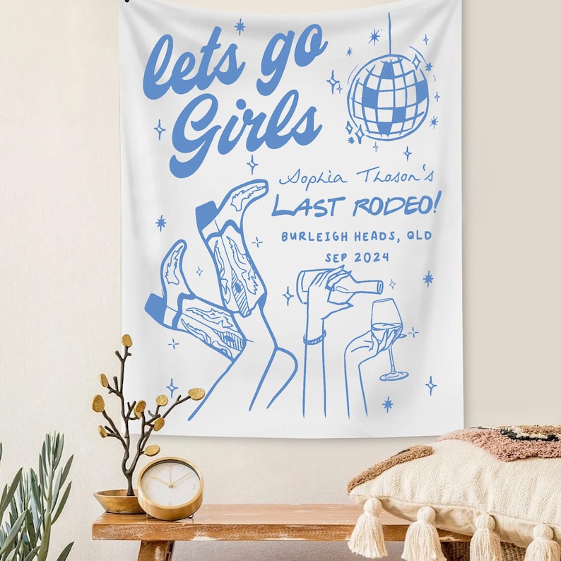 May include: White tapestry with blue text that reads "let's go girls" and "Sophia Thason's Last Rodeo!" with a disco ball graphic and a drawing of two women wearing cowboy boots and holding a bottle of wine and a glass of wine.