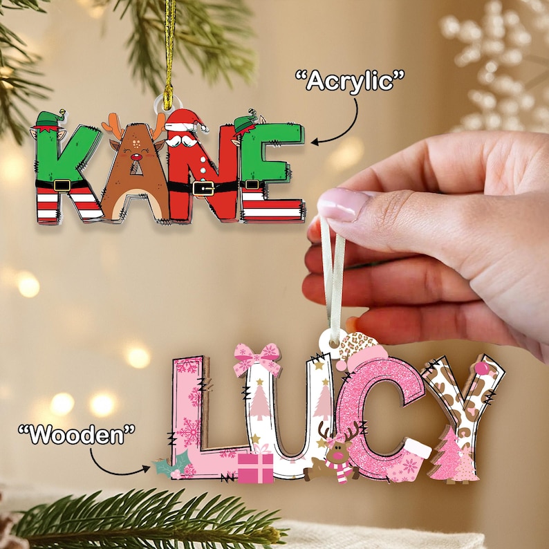May include: Two Christmas ornaments spelling out names. One is acrylic and spells "KANE" with festive designs. The other is wooden and spells "LUCY" with pink and white patterns, including trees and bows. Both have hanging ribbons.