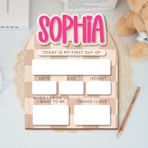 Op de afbeelding: Een houten bord met de naam "SOPHIA" in roze letters. Het bord heeft vakken om informatie over de eerste dag van een kind vast te leggen, waaronder datum, leeftijd en lengte. Het bevat ook vakken voor "Als ik groot ben wil ik zijn" en "Dingen die ik leuk vind".