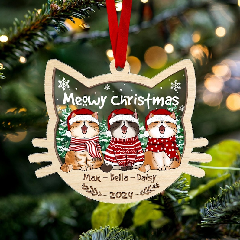 May include: Wooden cat-shaped Christmas ornament with the text "Meowy Christmas." The ornament features three cartoon cats wearing Santa hats and scarves. The names "Max - Bella - Daisy" and the year "2024" are also included. A red ribbon is attached for hanging.