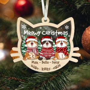 May include: Wooden cat-shaped Christmas ornament with three cartoon cats wearing Santa hats and scarves. The ornament says "Meowy Christmas" and has the names "Max - Bella - Daisy" and the year "2025" on it. The ornament hangs from a white ribbon.