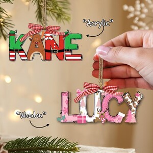 May include: Two Christmas ornaments spelling "KANE" and "LUCY". The "KANE" ornament is green, brown, and red with a plaid bow. The "LUCY" ornament is pink and white with a plaid bow. Both are hanging from twine.
