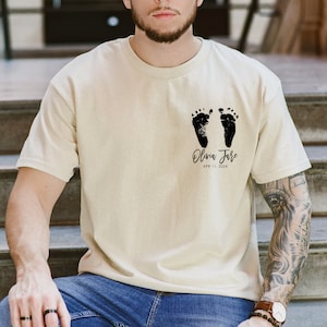 May include: A beige t-shirt with a black imprint of two baby footprints and the text "Olivia Jane APR 11, 2024".