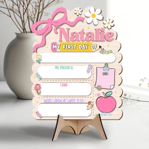 May include: A wooden sign for "Natalie" with the text "MY FIRST DAY OF". It features a pink bow, daisies, and a pink apple. Includes fill-in sections for "MY TEACHER IS", "I LOVE", and "WHEN I GROW UP I WANT TO BE". The sign is supported by a wooden stand.