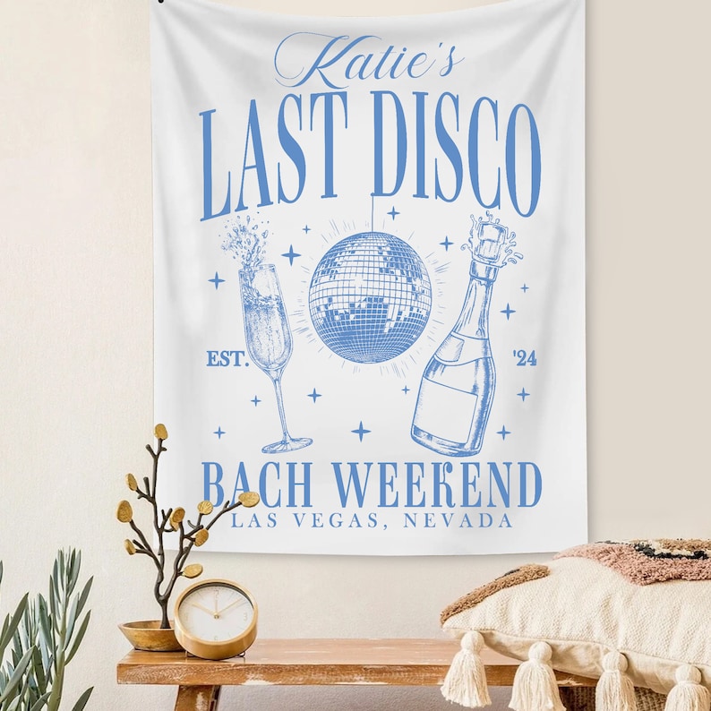 May include: White tapestry with blue text that reads "Katie's Last Disco Est. '24 Bach Weekend Las Vegas, Nevada". The tapestry also features a blue illustration of a champagne flute, a disco ball, and a bottle of champagne.
