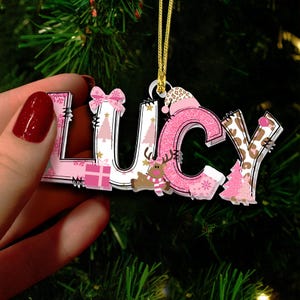 May include: A Christmas ornament spelling "LUCY" in pink, white, and brown. The letters are decorated with Christmas-themed patterns, including snowflakes, trees, and a reindeer. The "C" is glittery pink, and the "Y" has a leopard print.