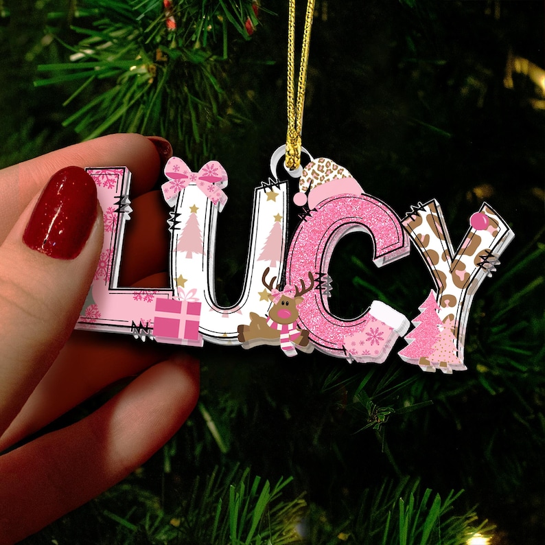May include: A Christmas ornament spelling "LUCY" in pink, white, and brown. The letters are decorated with Christmas-themed patterns, including snowflakes, trees, and a reindeer. The "C" is glittery pink, and the "Y" has a leopard print.