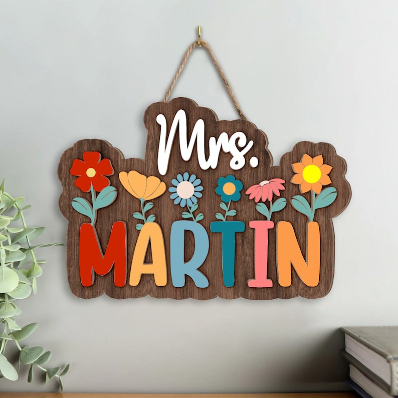 May include: A wooden sign with the text "Mrs. MARTIN" in colorful letters. The sign is decorated with colorful flowers and has a rope for hanging.