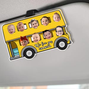 May include: A yellow school bus-shaped car visor ornament featuring the faces of several children. The bus has black wheels and the text "We love you Grandpa". The ornament is designed to clip onto a car visor.