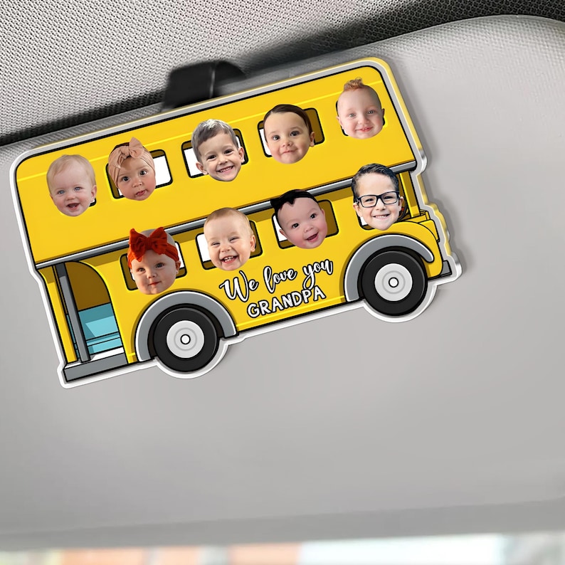 May include: A yellow school bus-shaped car visor ornament featuring the faces of several children. The bus has black wheels and the text "We love you Grandpa". The ornament is designed to clip onto a car visor.
