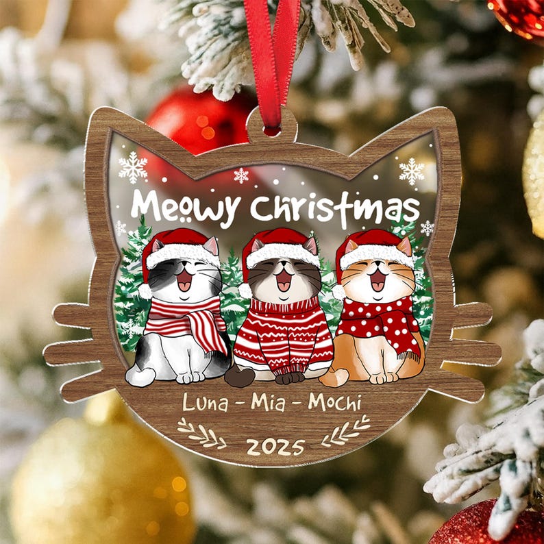 May include: Wooden Christmas ornament shaped like a cat's face, featuring three cartoon cats wearing Santa hats and scarves. The ornament says "Meowy Christmas" and includes the names "Luna - Mia - Mochi" and the year 2025.