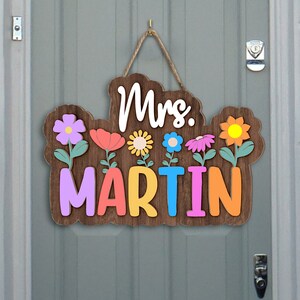 May include: A wooden door hanger with the text "Mrs. MARTIN" in colorful letters. The letters are surrounded by colorful flowers with green stems.