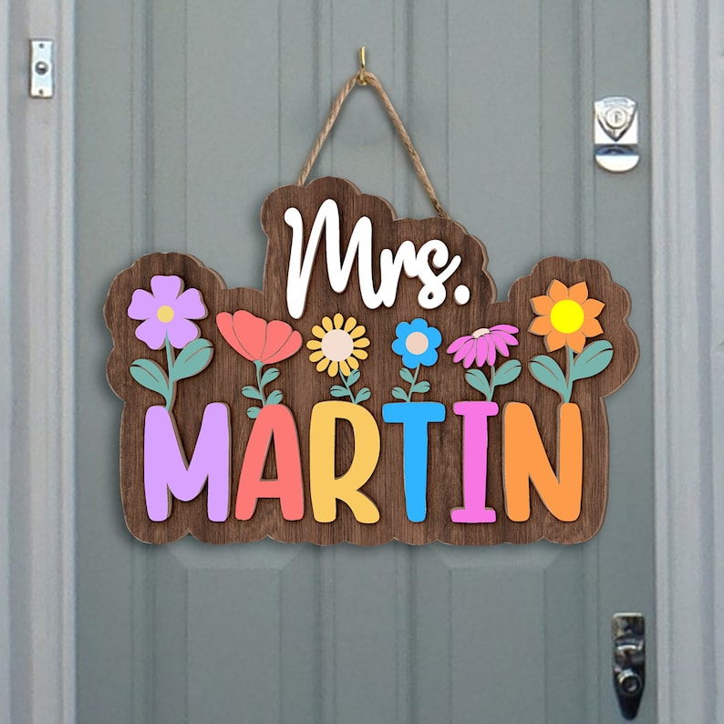 May include: A wooden door hanger with the text "Mrs. MARTIN" in colorful letters. The letters are surrounded by colorful flowers with green stems.