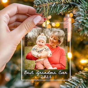 May include: Clear, square Christmas ornament with a photo of a baby and a woman. The baby wears a hat and sweater. The woman wears glasses and a red shirt. Text on the ornament reads "Best Grandma Ever 2025". A gold string hangs from the top.