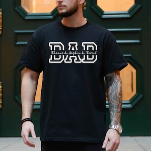 K&ouml;nnte beinhalten: Schwarzes T-Shirt mit wei&szlig;em Text, der "DAD" in einer College-Schriftart lautet. Unter dem Text steht "Thomas & Sophia & David" in einer kursiven Schrift.