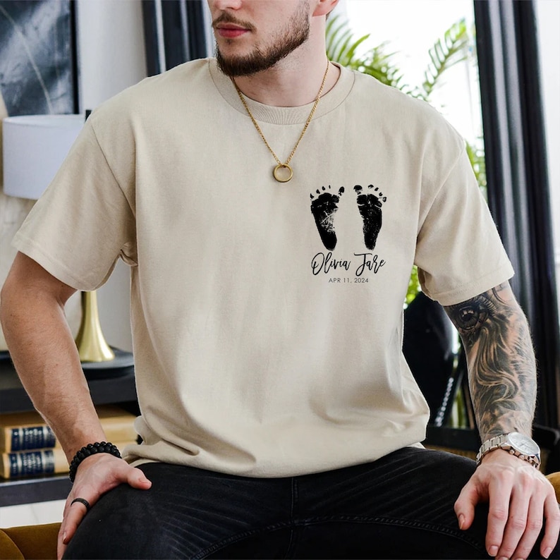 May include: A beige t-shirt with a black imprint of two baby footprints and the text "Olivia Jane APR 11, 2024".