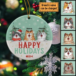 May include: A round, white ceramic ornament with a green background and a festive Christmas scene. Three cats with antlers are peeking over a banner that reads "Happy Holidays". The ornament is decorated with snowflakes and a fishbone.