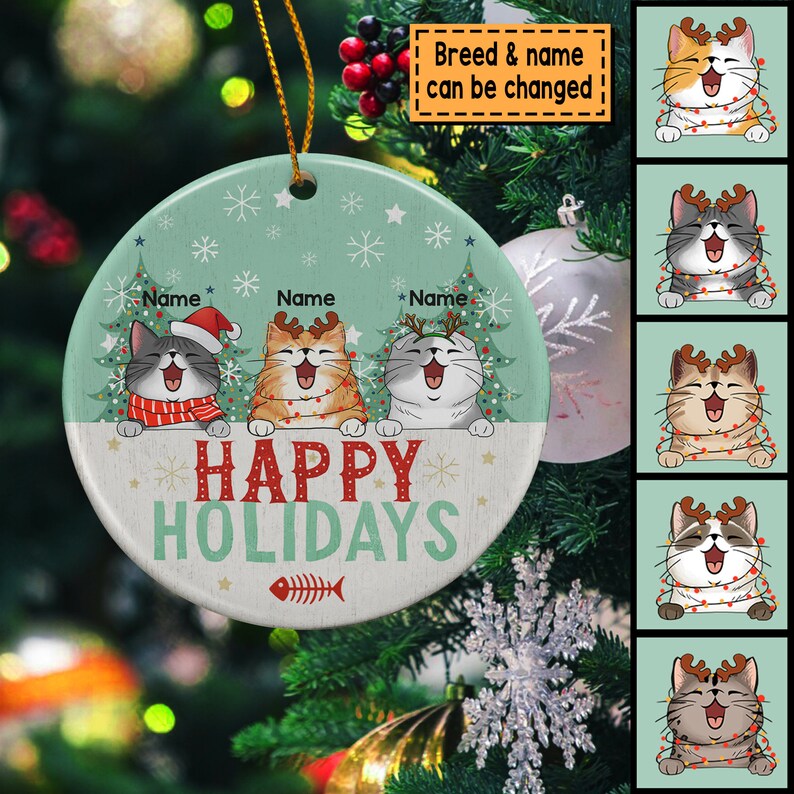 May include: A round, white ceramic ornament with a green background and a festive Christmas scene. Three cats with antlers are peeking over a banner that reads "Happy Holidays". The ornament is decorated with snowflakes and a fishbone.