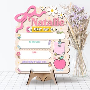 May include: A wooden first day of school sign, personalised with the name "Natalie" in pink. The sign features a bow, daisies, and spaces to record details such as teacher's name, date, and age. Includes a pencil, apple, and other decorative elements.