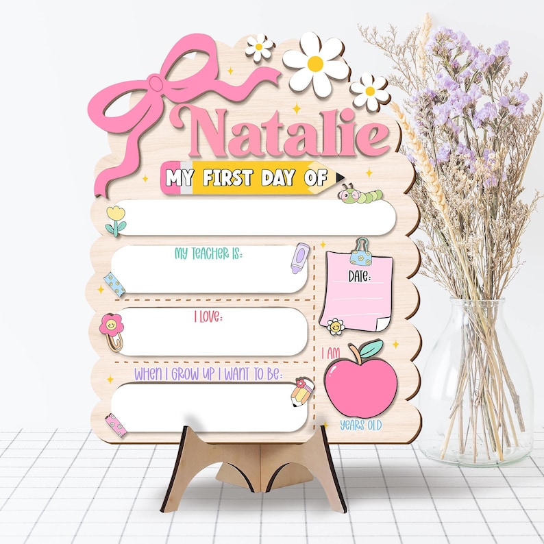 May include: A wooden first day of school sign, personalised with the name "Natalie" in pink. The sign features a bow, daisies, and spaces to record details such as teacher's name, date, and age. Includes a pencil, apple, and other decorative elements.
