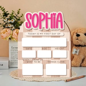 Op de afbeelding: Houten bord met de naam "SOPHIA" in roze letters. Het bord heeft vakken om "TODAY IS MY FIRST DAY OF", "DATE", "AGE", "HEIGHT", "WHEN I GROW UP I WANT TO BE" en "THINGS I LOVE" in te schrijven.