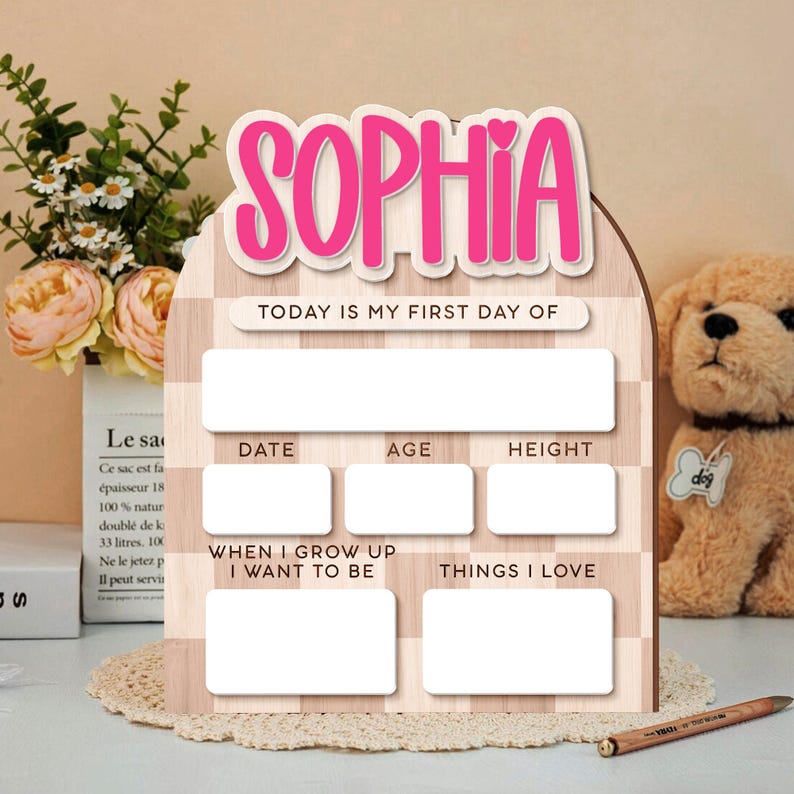 Op de afbeelding: Houten bord met de naam "SOPHIA" in roze letters. Het bord heeft vakken om "TODAY IS MY FIRST DAY OF", "DATE", "AGE", "HEIGHT", "WHEN I GROW UP I WANT TO BE" en "THINGS I LOVE" in te schrijven.