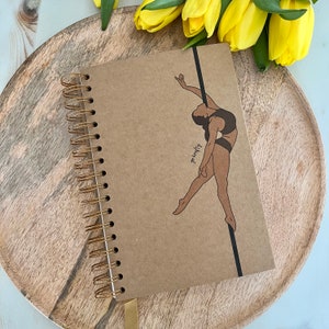 Pode incluir: Um caderno com espiral e capa de papel kraft. Apresenta uma ilustração castanha de uma pessoa a dançar numa barra. O caderno tem encadernação dourada e fecho de banda elástica preta. O texto "go krafty" está impresso.