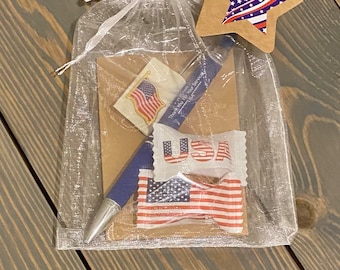 May include: A clear organza gift bag filled with patriotic themed items, including a pen, candy, and a sticker. The bag has a brown star-shaped tag with a red, white, and blue American flag design. The text on the tag reads "Veteran Gift Bag - all ready for gifting!"